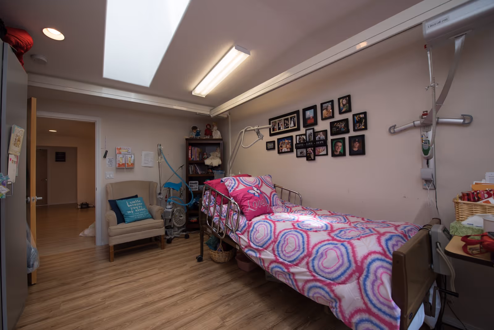 A small patient bedroom with a hospital-style bed dressed in bright pink patterned bedding, medical equipment, an armchair, a bookshelf and a photo collage on the wall beneath a skylight.
