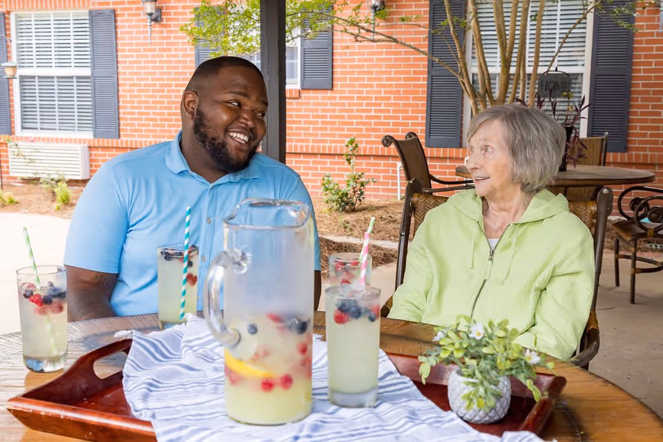 An elderly woman and a man sitting at an outdoor table under a covered patio, smiling and talking. On the table is a pitcher and glasses of lemonade with berries and striped straws, along with a small potted plant. The background shows a brick building with windows and some greenery.