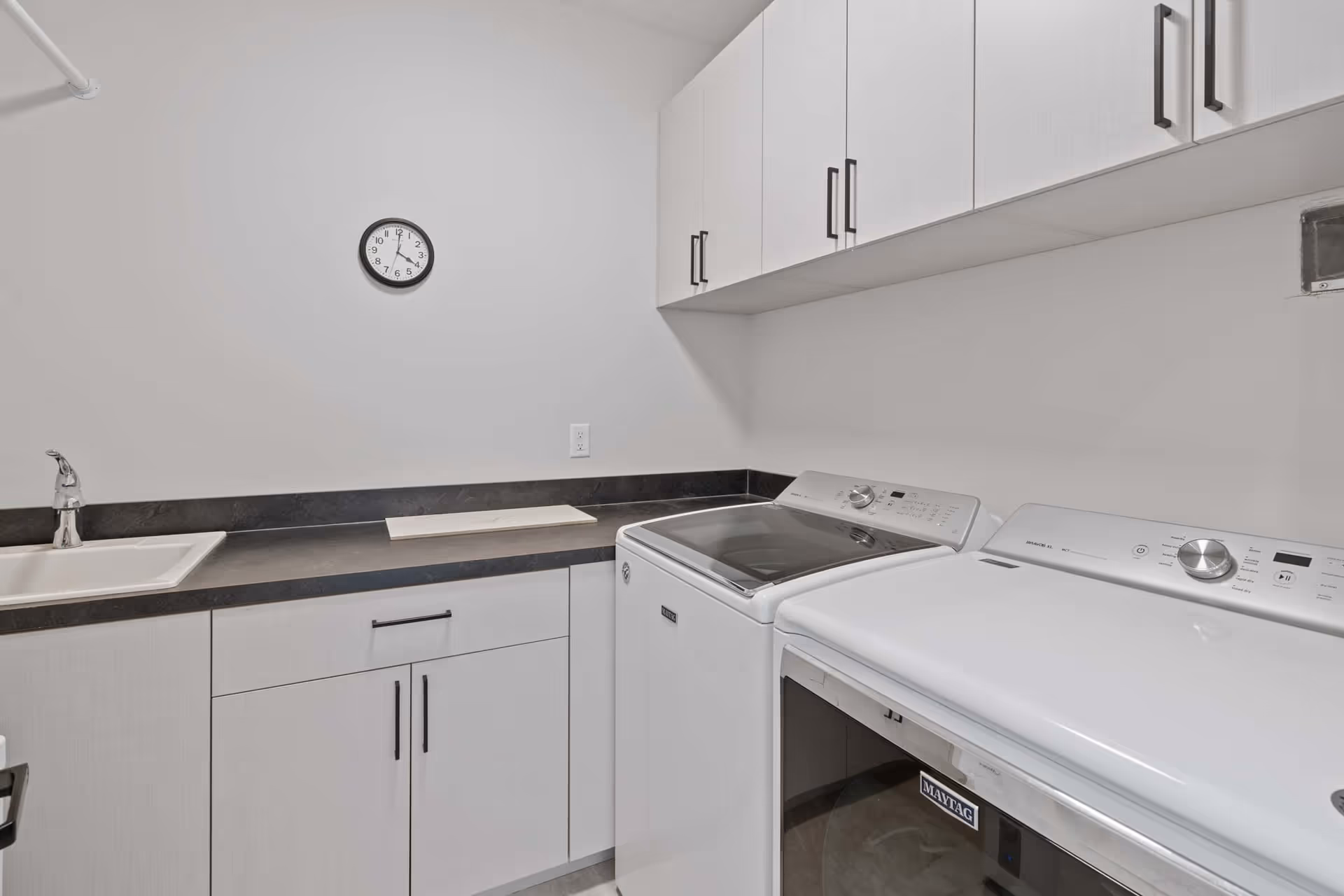 Laundry room with a white Maytag washing machine and dryer side by side, white cabinets above and below a black countertop, a white sink with a silver faucet, and a wall clock showing the time as 6:25.