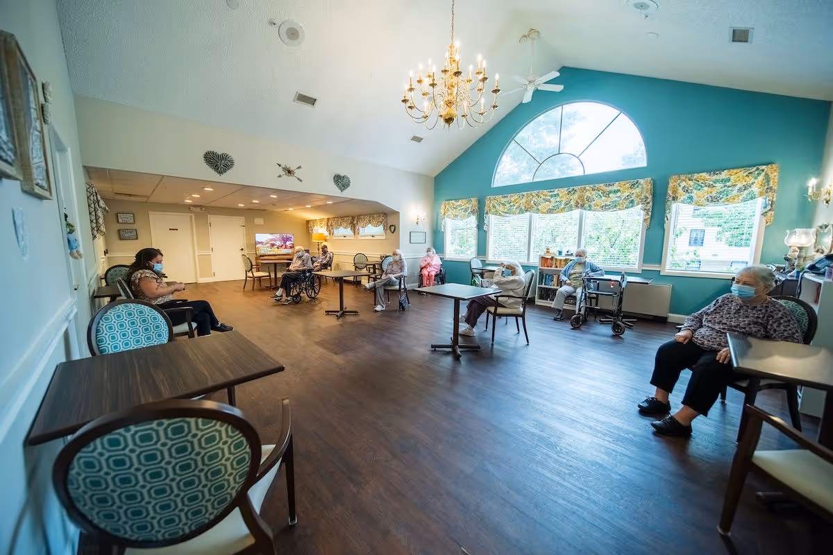 A spacious common room in a senior living facility with several elderly residents sitting apart at tables and chairs, all wearing face masks. The room has large windows with floral valances, a high ceiling with a chandelier, and light blue and white walls. The floor is wooden, and there are decorative wall hangings and a television in the background.