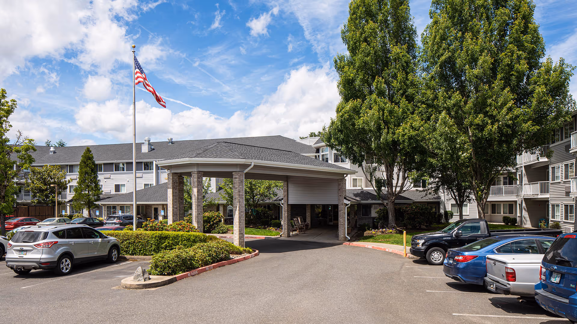 Exterior view of Holiday Rock Creek senior living facility showing the main entrance with a covered drop-off area, surrounded by parked cars, green trees, and a flagpole with an American flag against a partly cloudy blue sky.
