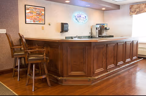 Interior view of a wooden bar counter with two high chairs in front. Behind the counter, there is a soda fountain machine and a neon sign that reads 'ICE CREAM SHOPPE'. The room has wooden flooring, beige walls, and a framed picture on the wall.