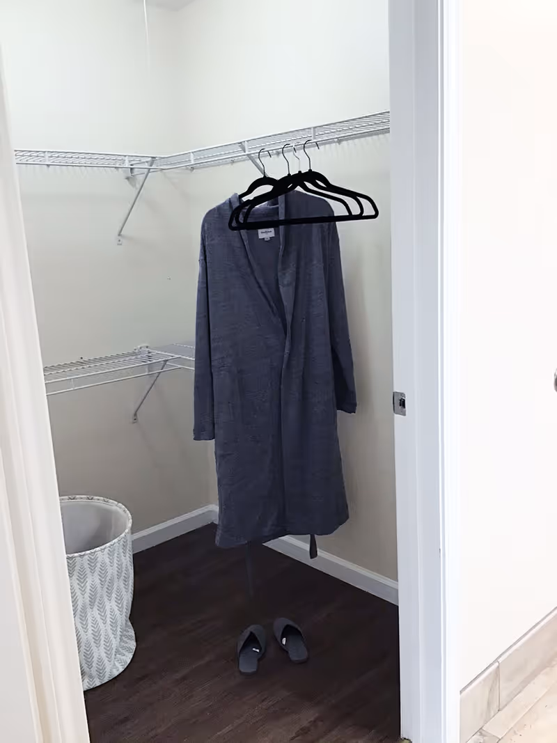 A small walk-in closet with a gray robe hanging on black hangers, a laundry basket to the side and a pair of slippers on a dark wood floor.