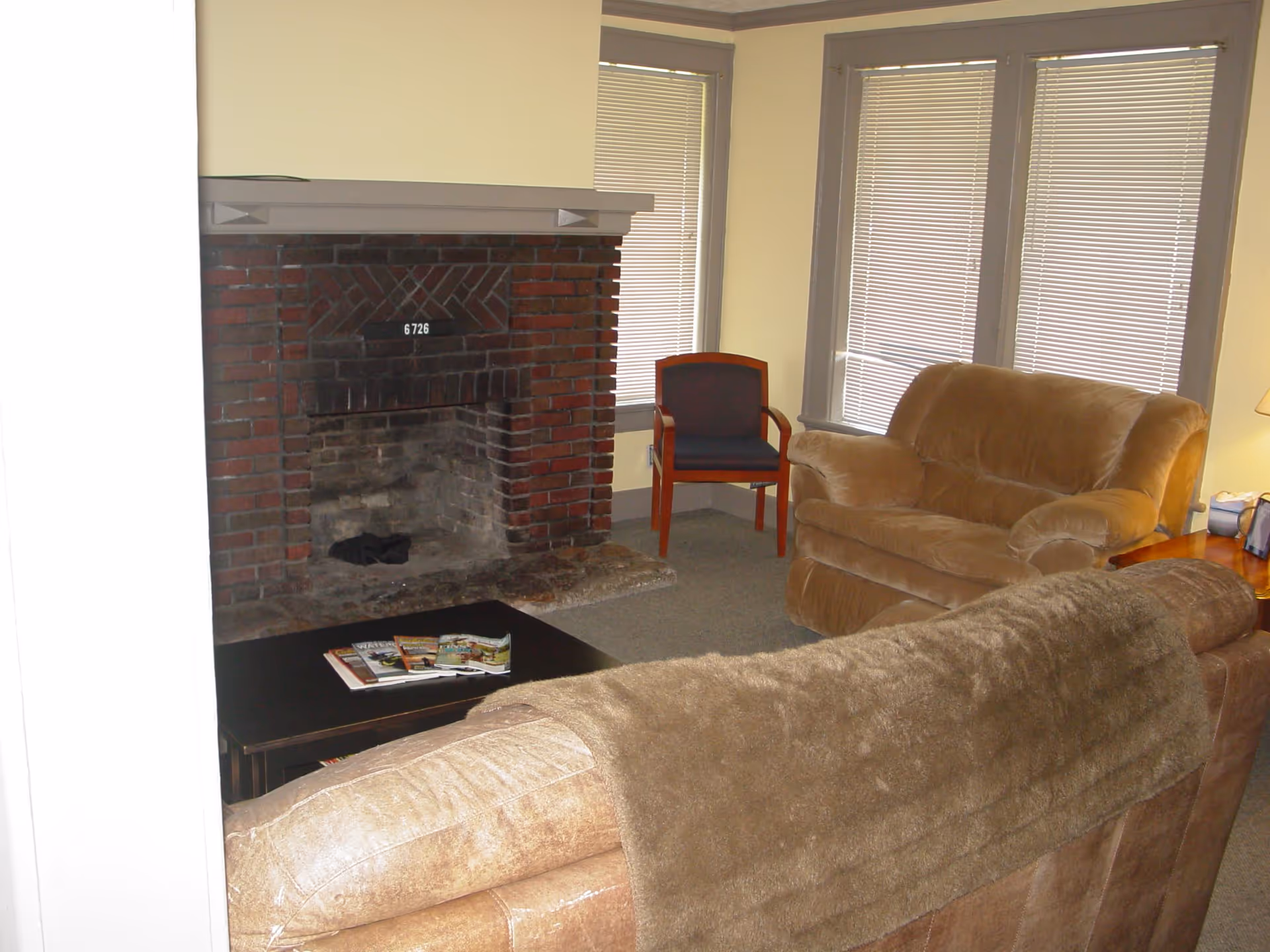 A cozy living room with a brown leather sofa covered partially by a brown blanket, a matching armchair, a wooden chair with dark blue upholstery, a dark coffee table with magazines on it, and a brick fireplace with a number plaque reading 6726. The room has three windows with closed blinds and light yellow walls.