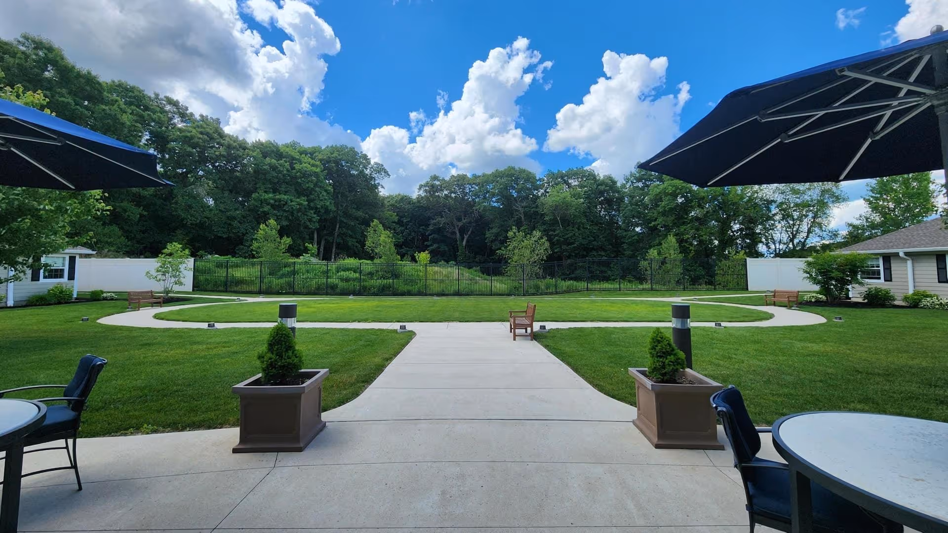 Outdoor patio area with round tables and chairs under large black umbrellas, overlooking a well-maintained lawn with a paved walkway, small trees, and benches. The background features a dense tree line under a partly cloudy blue sky.