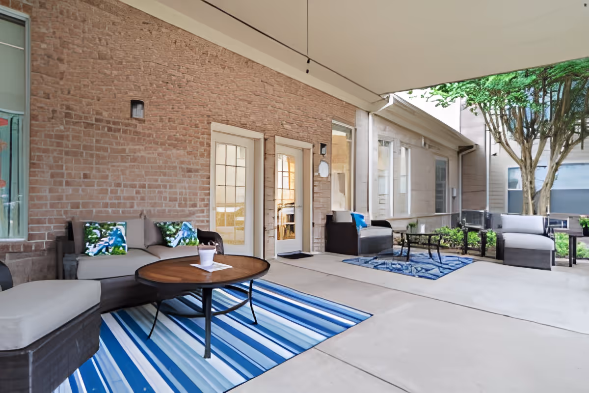 Covered outdoor patio area with cushioned seating, coffee tables, and blue striped rugs. The space is adjacent to a brick wall with glass-paneled doors and windows, and there are trees and greenery visible in the background.