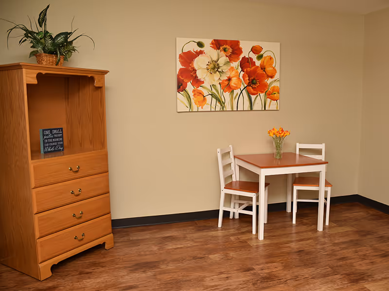 A small dining area with a wooden table and two white chairs with wooden seats. A vase with orange flowers is placed on the table. On the wall above the table is a painting of large orange and white flowers. To the left, there is a wooden cabinet with three drawers and an open shelf, decorated with a potted plant on top and a small framed sign inside the shelf. The room has wooden flooring and beige walls.