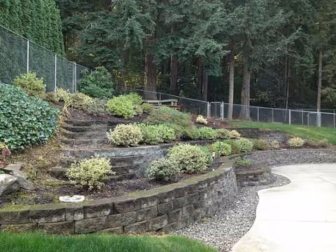Outdoor landscaped terrace with stone walls, shrubs, trees, a fence, and a concrete path.