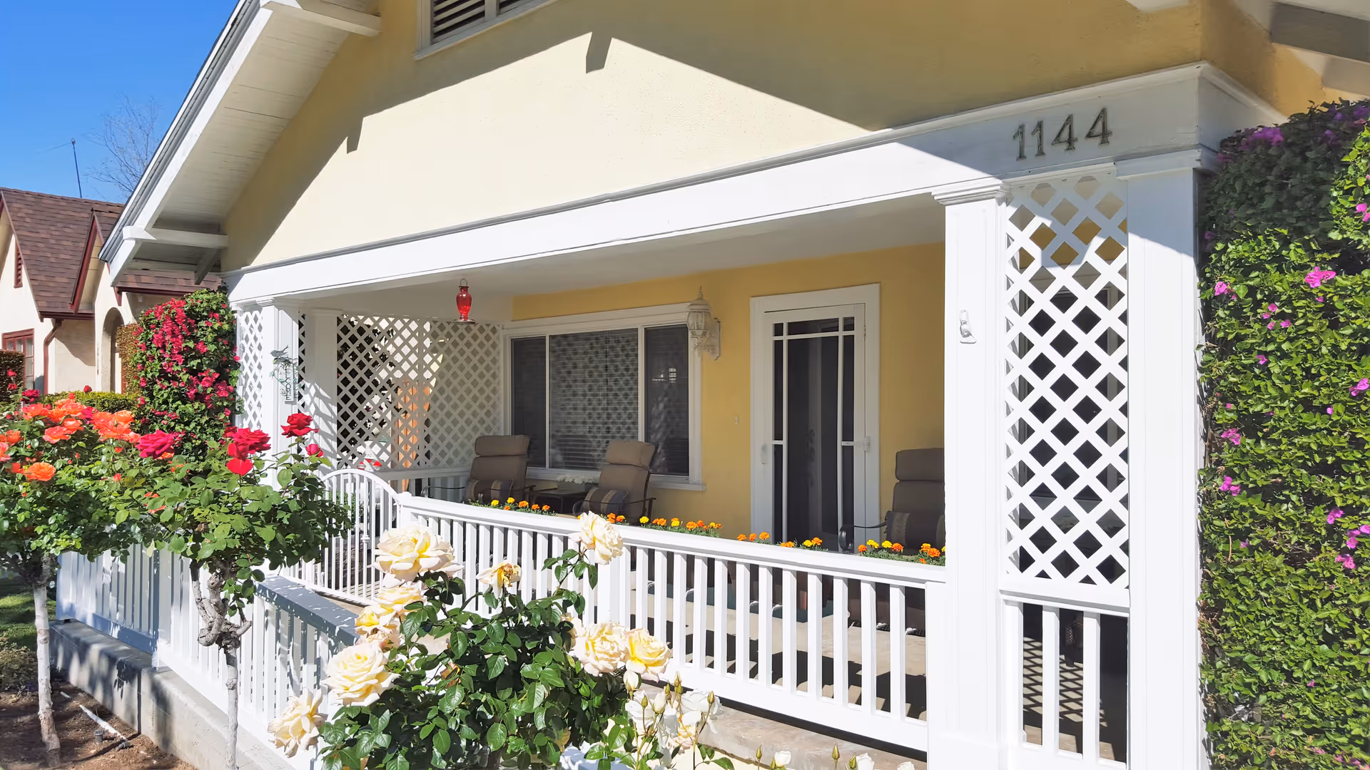 Front porch of a yellow house with white lattice columns and railing, featuring several cushioned chairs and flower boxes with orange flowers. The porch is surrounded by blooming rose bushes in red, orange, and pale yellow colors. The house number 1144 is visible on the upper right corner of the porch.