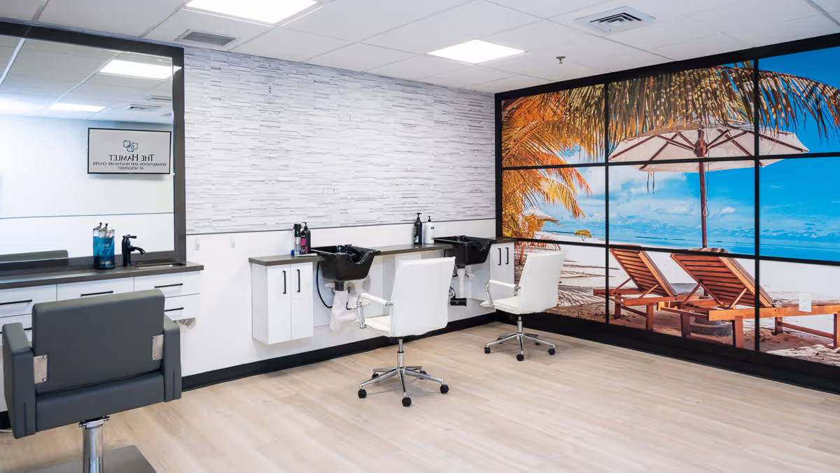 Interior view of a salon area in a healthcare facility featuring two white salon chairs in front of black wash basins, a large mirror on the left wall, and a large wall mural depicting a beach scene with lounge chairs and an umbrella on the right.