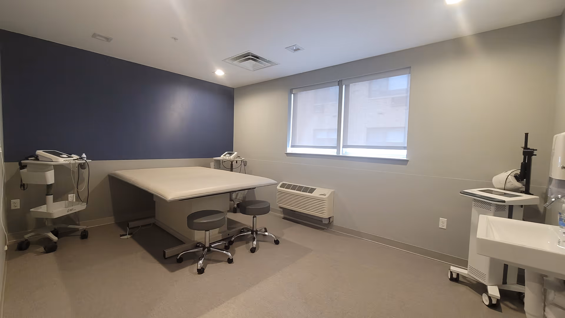 A clinical examination room with a large examination table in the center, two rolling stools, medical equipment on carts, a wall-mounted air conditioning unit below a window with blinds, and a sink on the right side. The walls are painted in neutral tones with one dark blue accent wall.