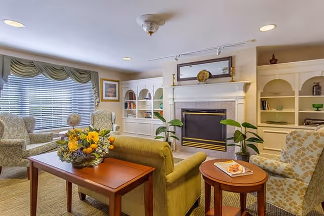 A cozy living room area with a green sofa, two patterned armchairs, a wooden coffee table with a floral arrangement, and a small round side table with a magazine. The room features a fireplace with a mirror and decorative items on the mantel, built-in white shelves with books and decor, and a window with green valance curtains.