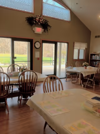Interior view of a senior living facility dining room with several tables covered with tablecloths and chairs around them. There are large windows and glass doors letting in natural light and showing a green outdoor area. The room has a high ceiling with a floral arrangement above the exit door and a clock on the wall.