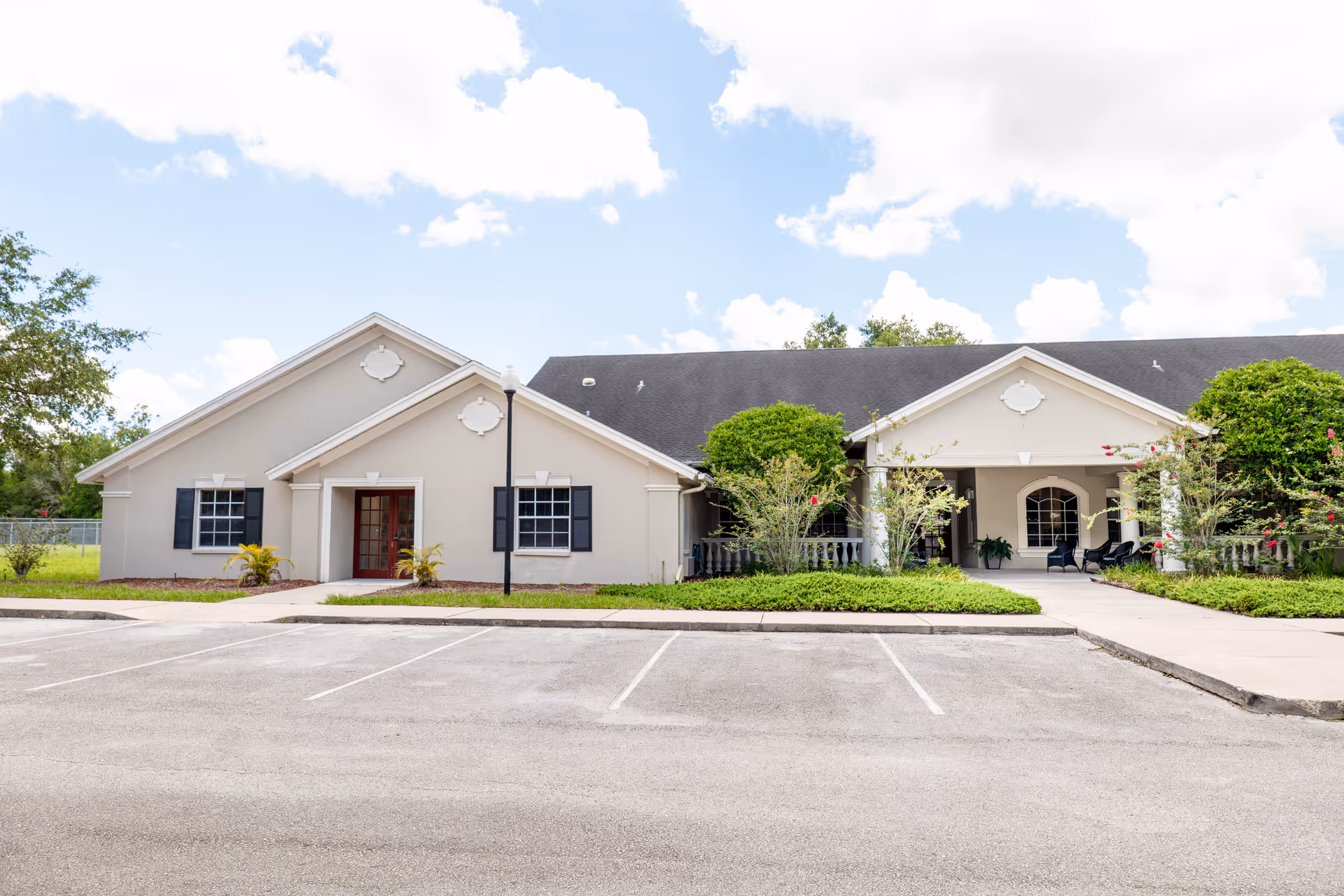 Exterior view of a single-story building with beige walls, black shutters, and a dark roof under a partly cloudy sky. There is a parking lot with empty spaces in front of the building, and landscaped greenery with bushes and small trees near the entrance.
