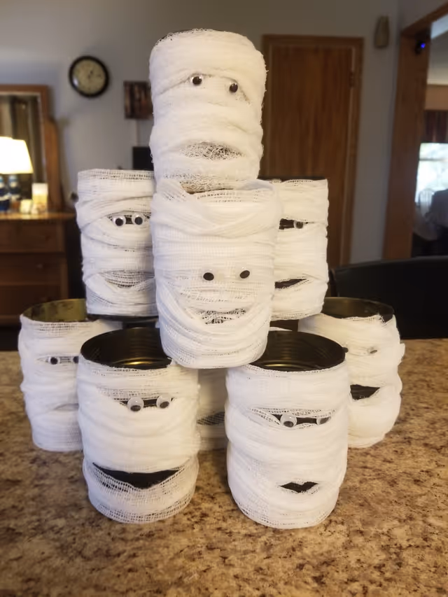 Tin cans wrapped in white gauze with googly eyes resembling mummies, stacked on a countertop in a home interior.