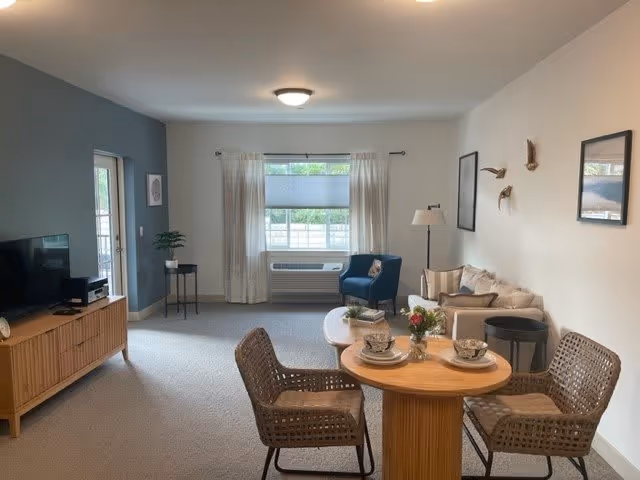 A cozy living room area in a senior living facility featuring a small round wooden table set with two cups and saucers, surrounded by two woven chairs. In the background, there is a beige sofa with cushions, a floor lamp, and wall decorations including framed pictures and bird sculptures. A window with light curtains and a window air conditioning unit is centered on the far wall. To the left, a wooden TV stand holds a flat-screen television and other electronic devices. The walls are painted white with one accent wall in blue-gray.