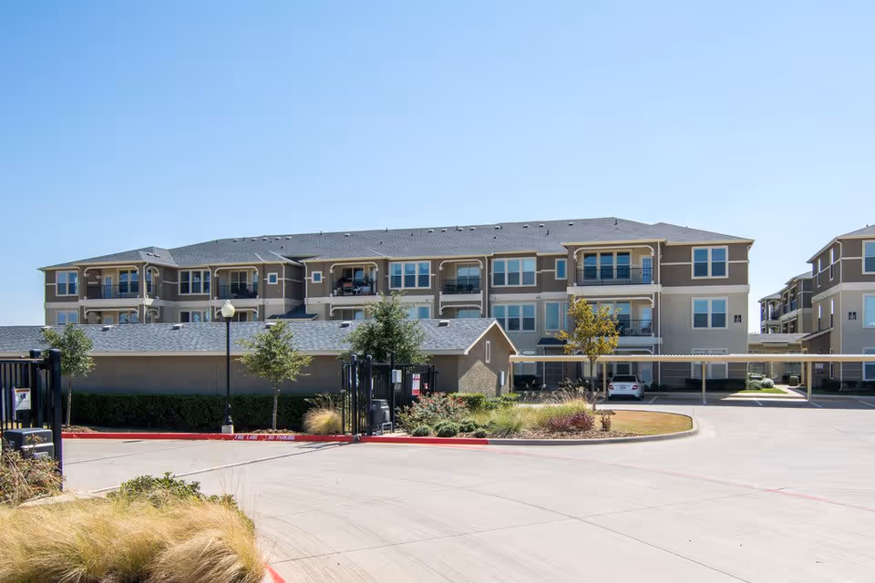 Exterior view of a multi-story residential building with balconies, a gated entrance, and a parking area with covered spaces under a clear blue sky.