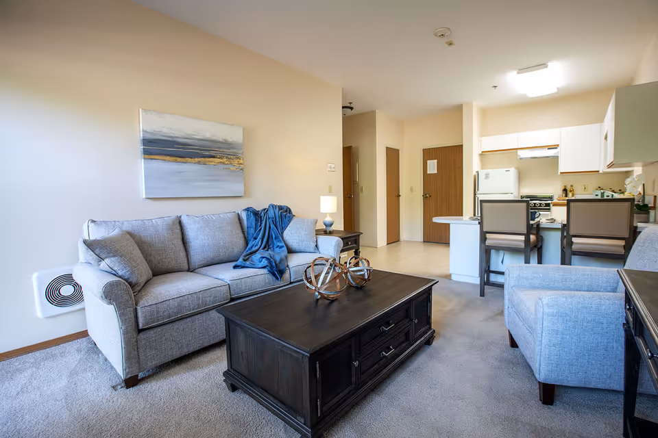 A cozy living room area with a light gray sofa and matching armchair, a dark wooden coffee table with decorative items, and a blue throw blanket on the sofa. The room has beige walls with a landscape painting above the sofa. In the background, there is a small kitchen area with white cabinets, a refrigerator, stove, and two chairs at a counter.