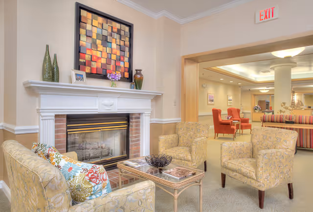 A cozy living room area in a senior living facility featuring a white fireplace with decorative vases and a colorful abstract painting above it. The room has two patterned armchairs and a matching loveseat with floral cushions arranged around a glass-top coffee table. In the background, there are additional seating areas with red armchairs and a striped sofa under soft lighting.