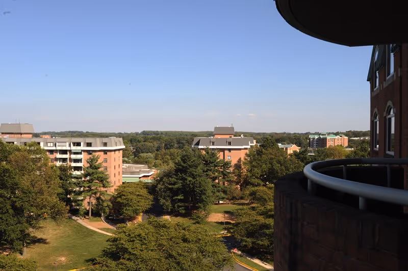 View from a balcony overlooking a senior living community with multiple brick buildings surrounded by trees and greenery under a clear blue sky.