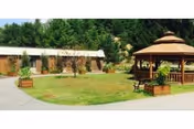 Outdoor view of a senior living facility featuring a grassy courtyard with a wooden gazebo, benches, potted plants, and a single-story brick building surrounded by trees.
