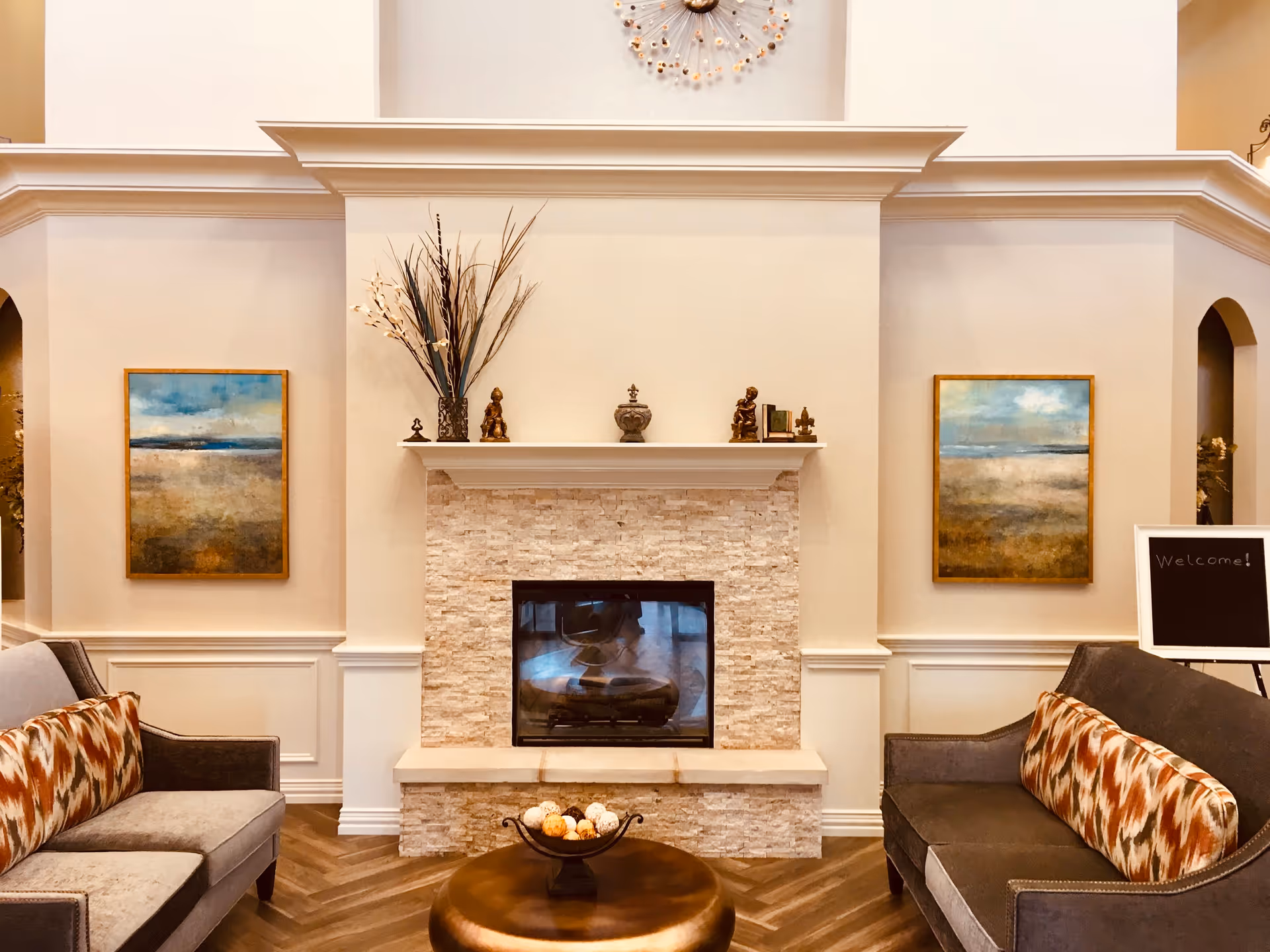 A cozy living room area with a stone fireplace in the center. On the mantel are decorative items including vases and small statues. Two framed landscape paintings hang on the walls on either side of the fireplace. There are two gray sofas facing each other, each with a patterned cushion. A round bronze coffee table with decorative balls is in front of the fireplace. A small chalkboard sign with the word 'Welcome!' is visible on the right side.