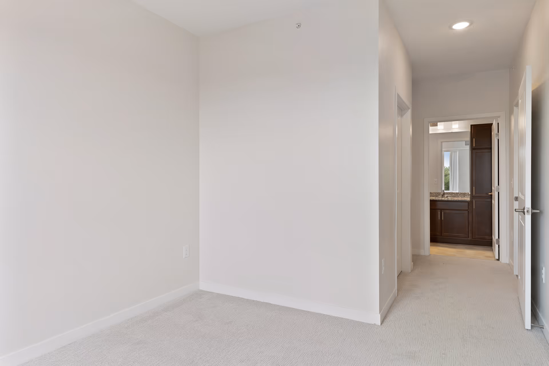Empty room with light beige carpet and white walls, leading to a hallway with open doors and a view into a bathroom with dark wood cabinets and a granite countertop.