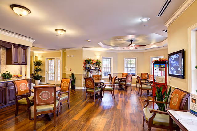 A bright and spacious common area with wooden flooring, multiple round tables surrounded by chairs with patterned cushions, a popcorn machine, and large windows letting in natural light. The room has a ceiling fan, recessed lighting, and decorative plants placed around the space.