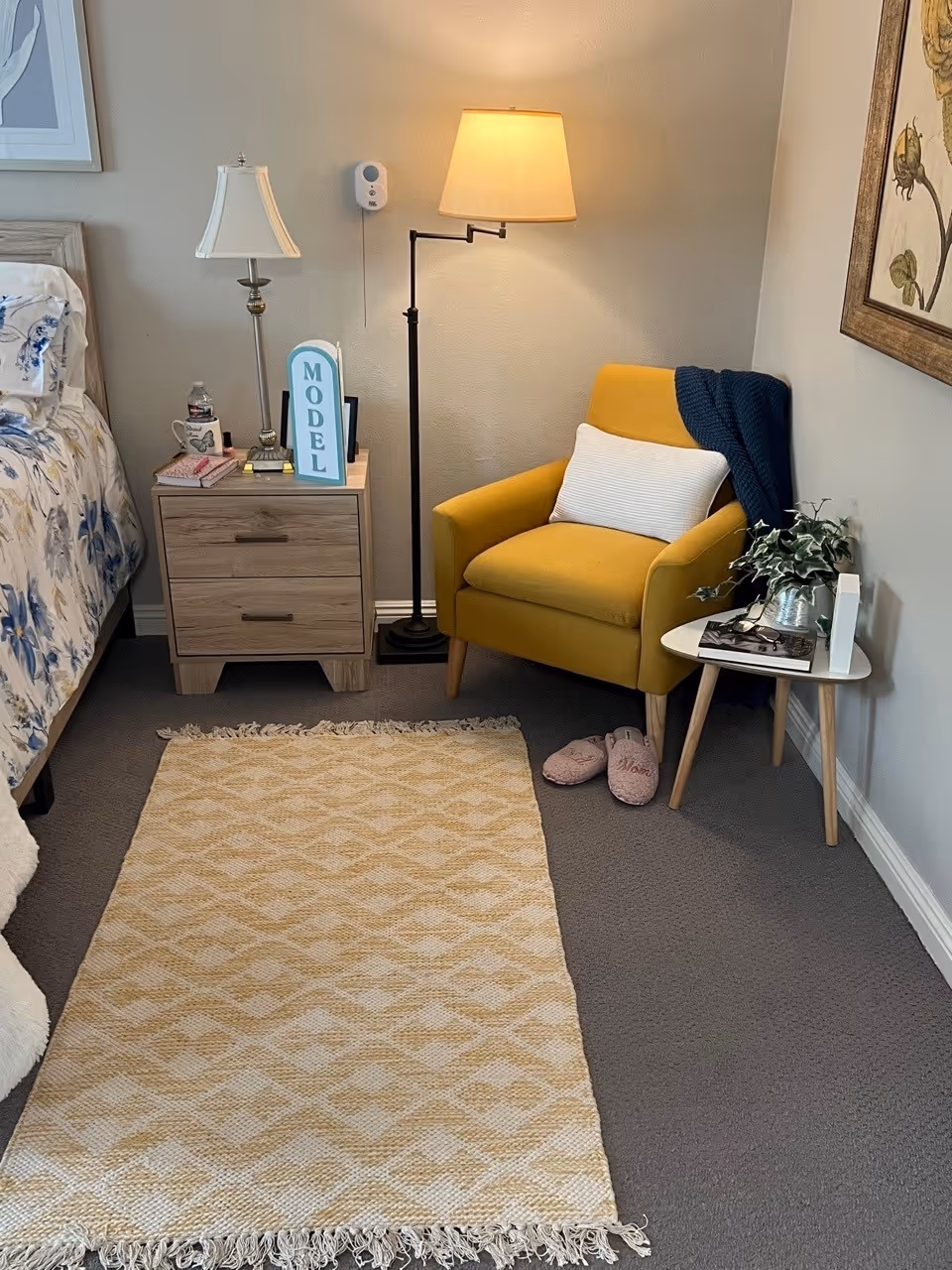 A cozy corner of a bedroom featuring a yellow armchair with a white pillow and a dark blue throw draped over it. Next to the chair is a small white side table with a plant, a pair of glasses, and a book. A pair of pink slippers is placed on the floor near the chair. To the left, there is a wooden nightstand with two drawers, a table lamp, a water bottle, a mug, and a sign that reads 'MODEL'. A floor lamp with a beige shade stands behind the armchair, and a beige patterned rug lies on the carpeted floor. Part of a bed with floral bedding is visible on the left side of the image.
