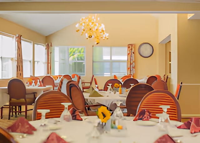 A dining room with multiple round tables covered with white tablecloths, set with glasses, napkins, and small flower centerpieces. The room has large windows with curtains, a chandelier hanging from the ceiling, and several wooden chairs around the tables.