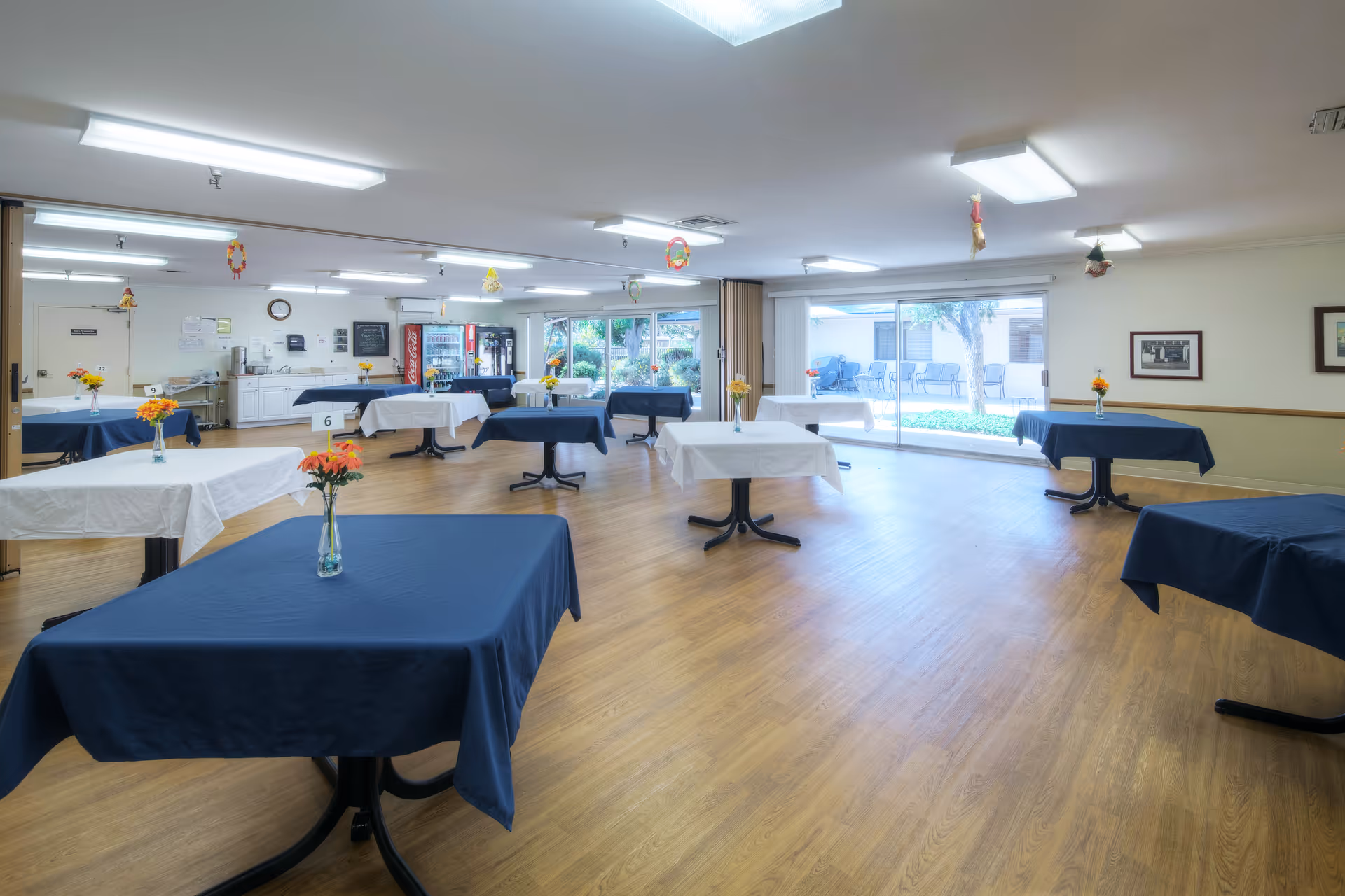 A spacious dining room with multiple tables covered in alternating white and navy blue tablecloths, each table adorned with a small vase of orange and yellow flowers. The room has wooden flooring, bright overhead lighting, and large sliding glass doors that open to an outdoor patio area with chairs and greenery.