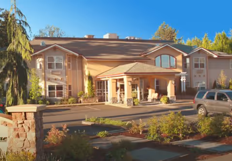 Exterior view of Hearthstone of Beaverton, a two-story building with a covered entrance, surrounded by landscaped greenery and a parked vehicle in front.