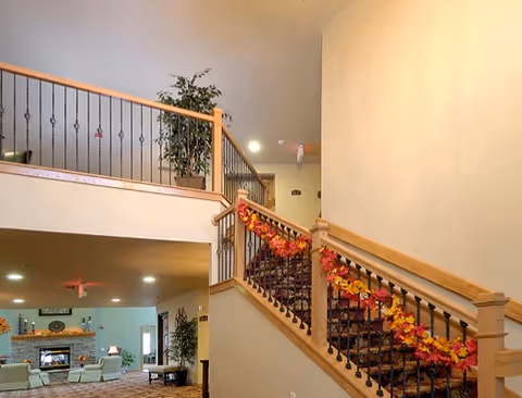 Interior view of a senior living facility showing a staircase decorated with autumn-themed garlands. The staircase has wooden handrails and black metal balusters. Below the staircase is a cozy living area with armchairs, a fireplace, and plants. The walls are painted in neutral tones and the space is well-lit with ceiling lights.
