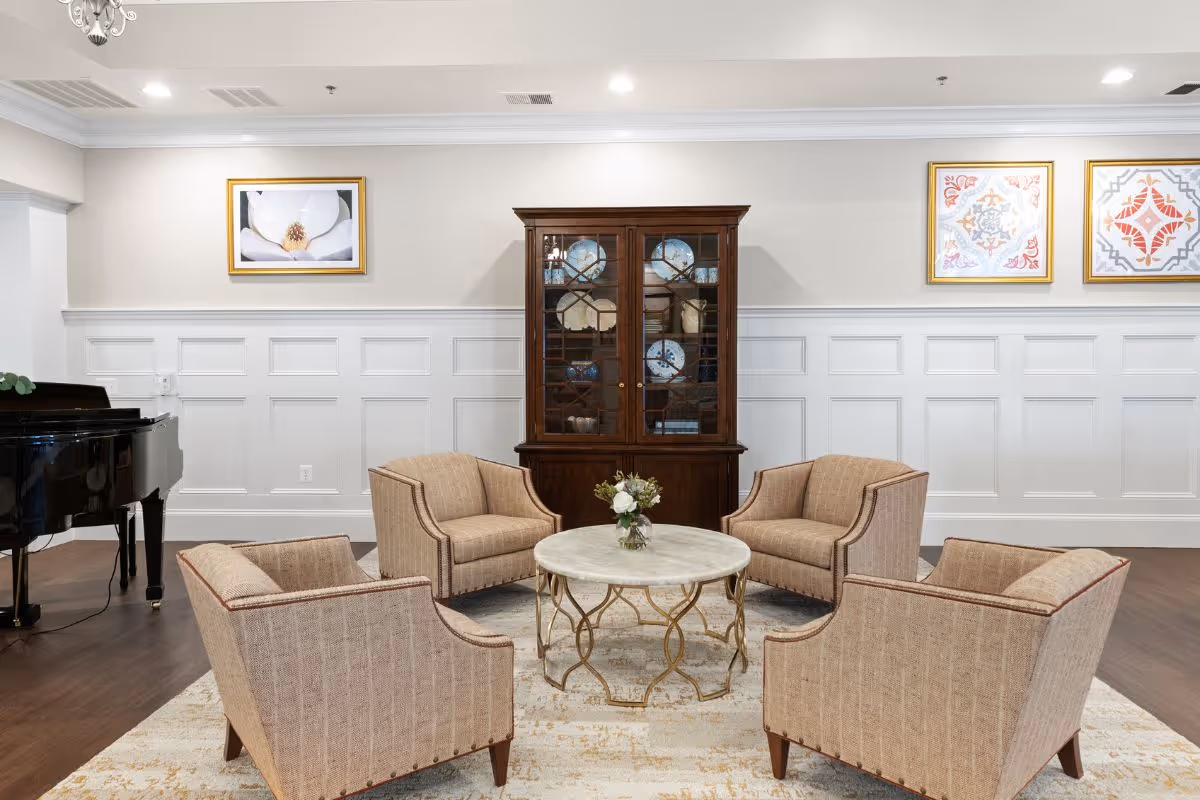 A cozy sitting area with four beige upholstered armchairs arranged around a round marble-top coffee table with a small floral arrangement. Behind the seating is a dark wooden cabinet displaying decorative plates. The room features white wainscoting on the walls, three framed artworks, and a black grand piano on the left side.