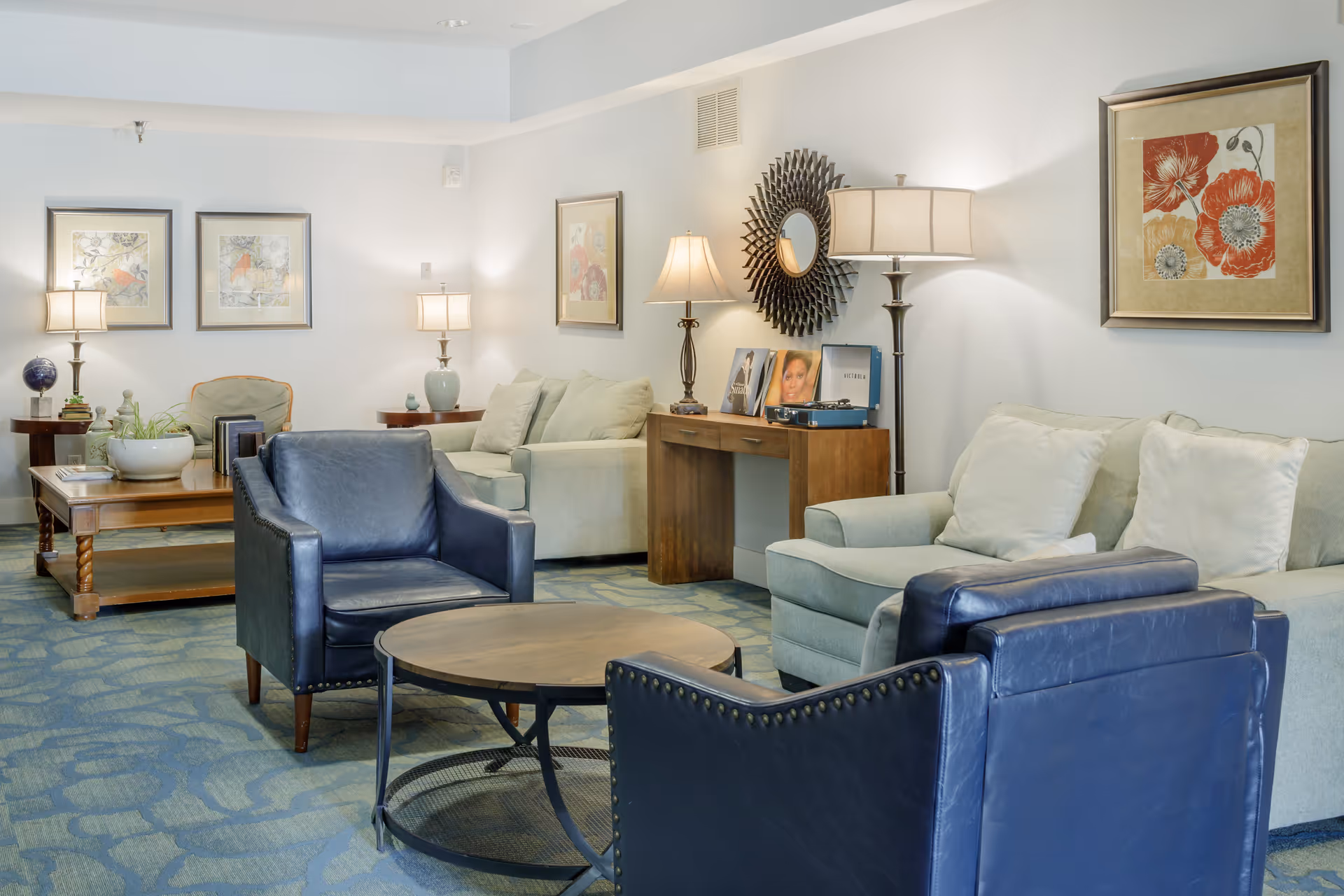 A cozy living room area with two light-colored sofas, two dark blue leather armchairs, a round wooden coffee table, and a rectangular wooden side table with lamps and decorative items. The walls are adorned with framed floral artwork and a decorative round mirror. The room has a soft blue patterned carpet and soft lighting from table and floor lamps.