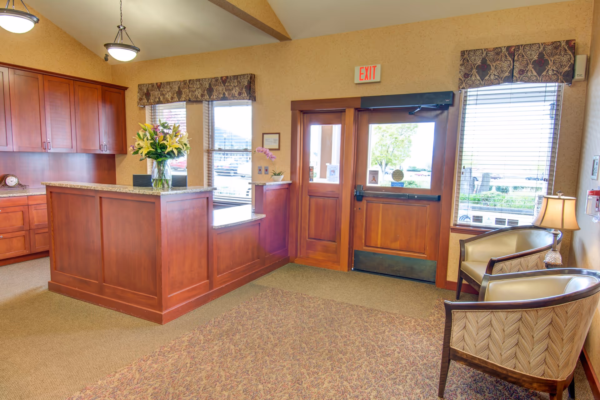 Reception area with a wooden front desk, two armchairs with patterned upholstery, a table lamp, and windows with blinds. There is an exit door with glass panels and an exit sign above it. The room has beige walls and carpeted floor with a patterned rug.