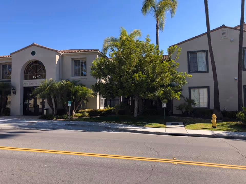 Exterior view of Silvergate San Marcos facility showing a two-story building with beige walls, tiled roof, several windows, palm trees, and a fire hydrant near the sidewalk under a clear blue sky.