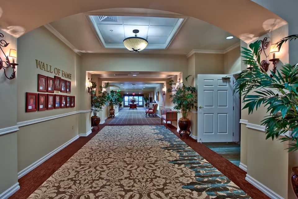 A well-lit hallway in a senior living facility with patterned carpet, potted plants along the walls, and a 'Wall of Fame' display featuring framed photos on the left wall. There are wall sconces providing warm lighting, a ceiling light fixture, and an open door on the right side leading to another room. At the far end, seating and a table with a floral arrangement are visible near large windows.