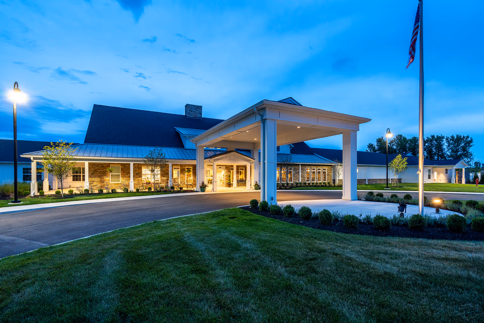 Front exterior of Walnut Crossing senior living facility at dusk with a covered entrance canopy, landscaped lawn, and flagpole.