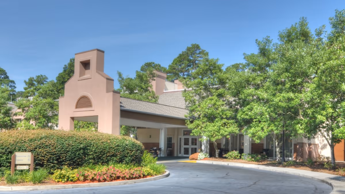 Exterior view of the entrance to Savannah Square senior living facility with a covered drop-off area, surrounded by green trees and landscaped bushes under a clear blue sky.