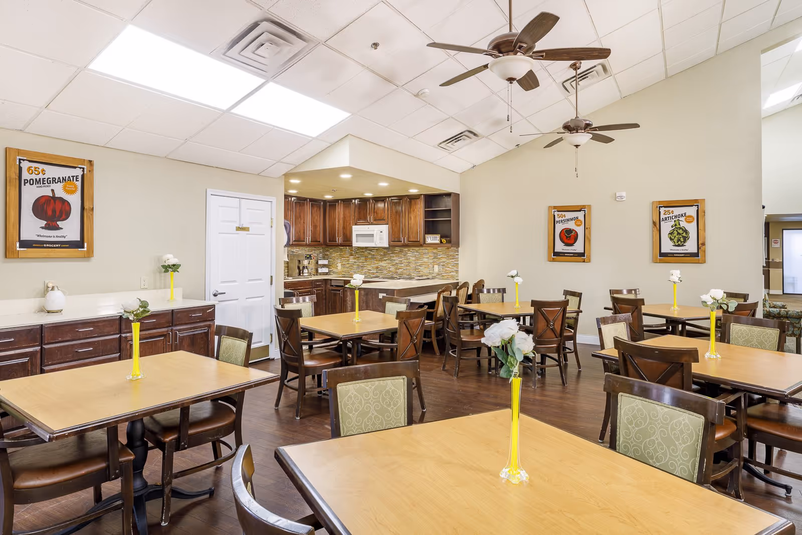 Dining area with multiple wooden tables and chairs, small vases with flowers, ceiling fans, and an open kitchen in the back.