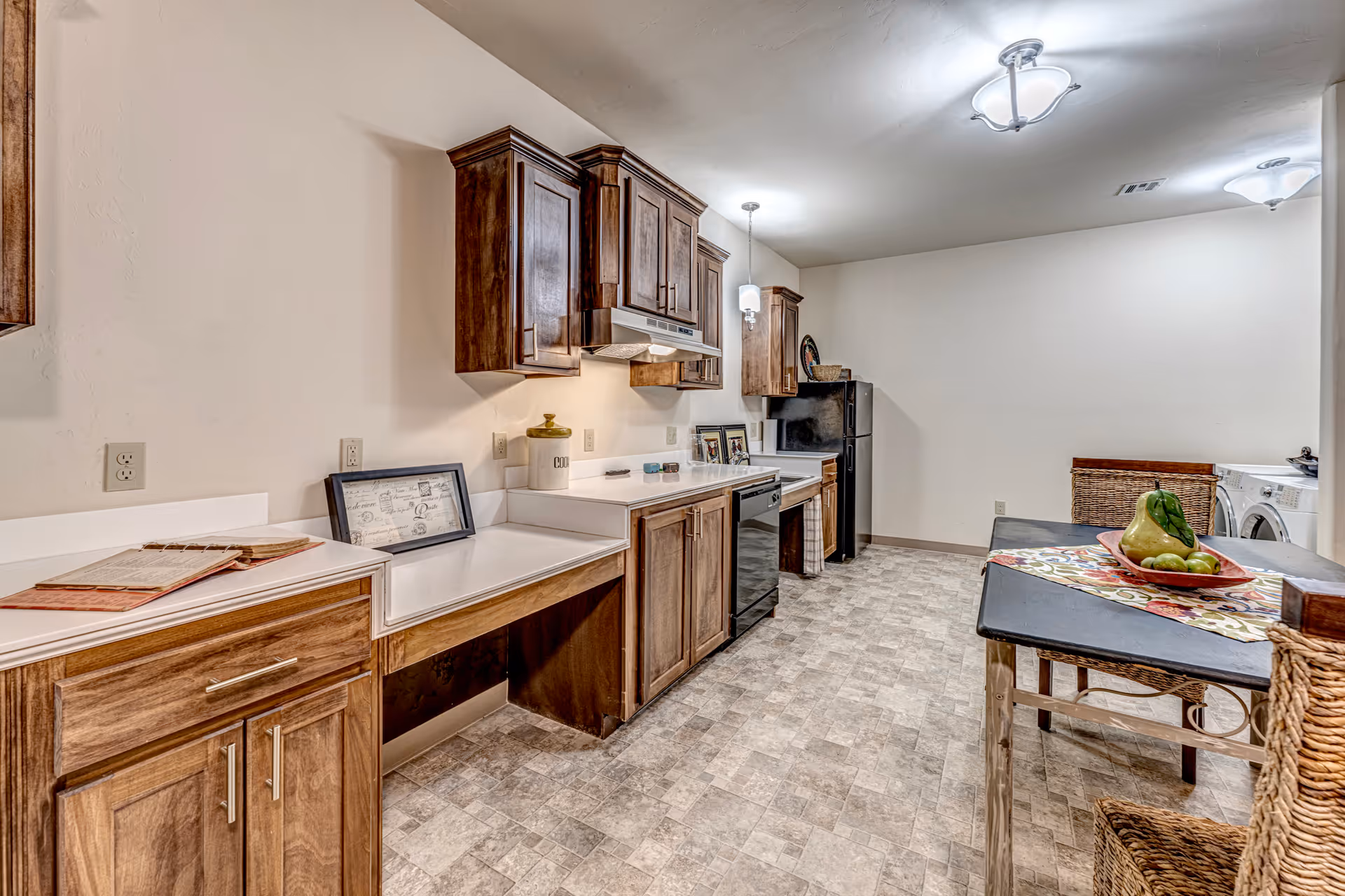A spacious kitchen area with wooden cabinets, white countertops, and modern appliances including a black refrigerator and dishwasher. There is a table with a colorful runner and a fruit bowl containing pears and apples. The room also features a washing machine and dryer in the background, with wicker chairs around the table and soft ceiling lights illuminating the space.