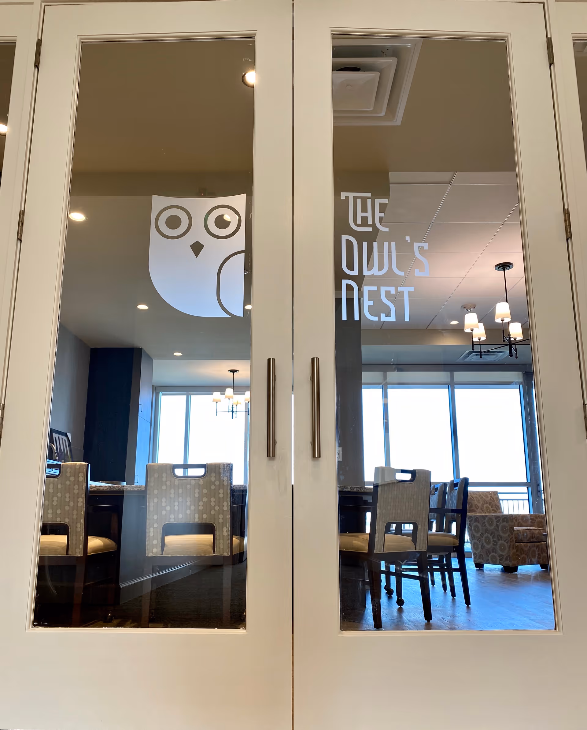 Glass double doors with an owl graphic and the words "The Owl's Nest" revealing a dining/lounge area with chairs and hanging lights.