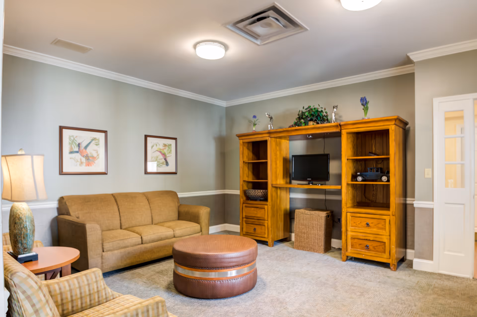 A cozy living room with a sofa, armchair, round ottoman, wooden entertainment center with a TV, and framed artwork on the wall.