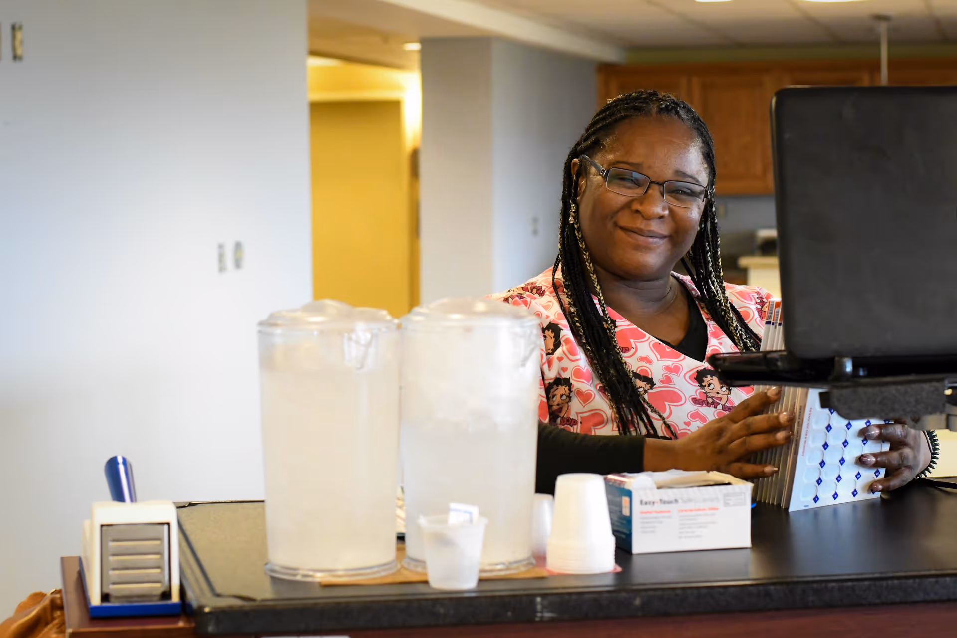 A smiling woman with braided hair and glasses stands behind a counter with two large pitchers of ice water, disposable cups, and a box of safety lancets. She is wearing a pink scrub top with heart and cartoon character patterns and is holding a file organizer. The background shows a light-colored wall and wooden cabinets.