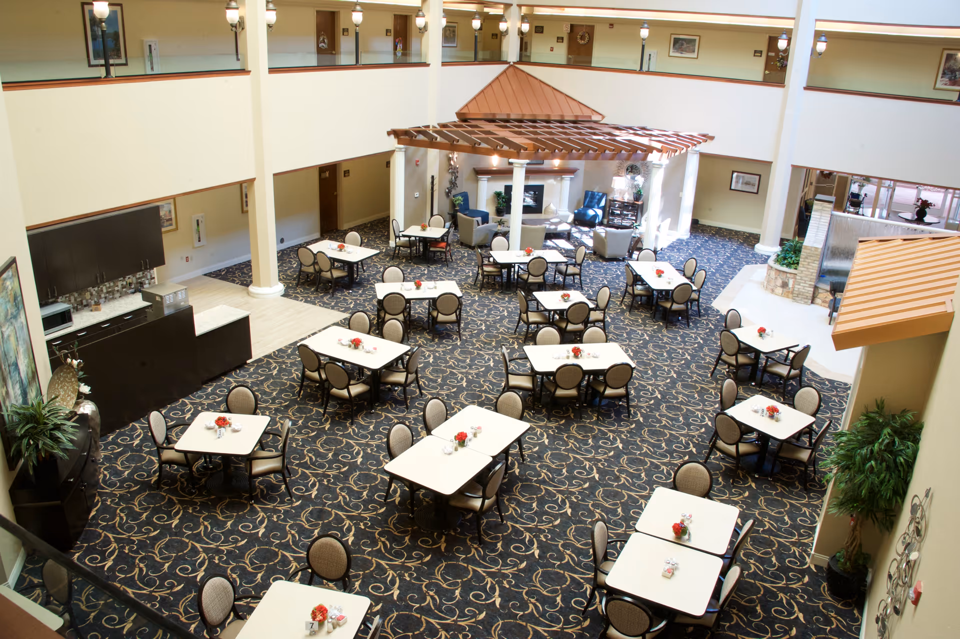 Spacious two-story atrium dining room with multiple square tables and chairs arranged around a central pergola and seating area.