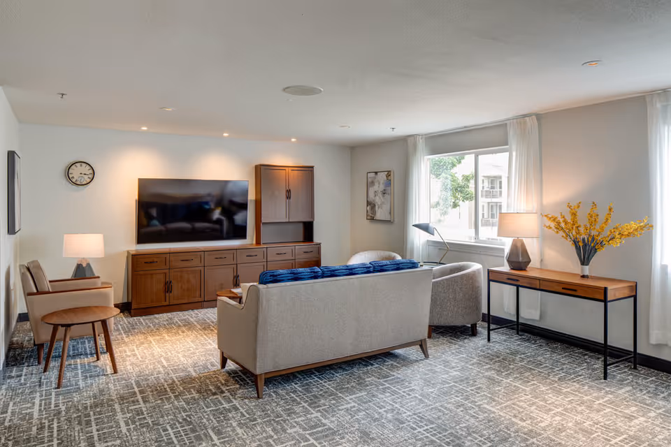 A bright and modern living room with a large flat-screen TV mounted on the wall above a wooden cabinet. The room features a beige sofa with a blue throw blanket, two beige armchairs, a wooden side table, a console table with a lamp and a vase of yellow flowers, and a large window with white curtains letting in natural light. The floor is carpeted with a patterned design.