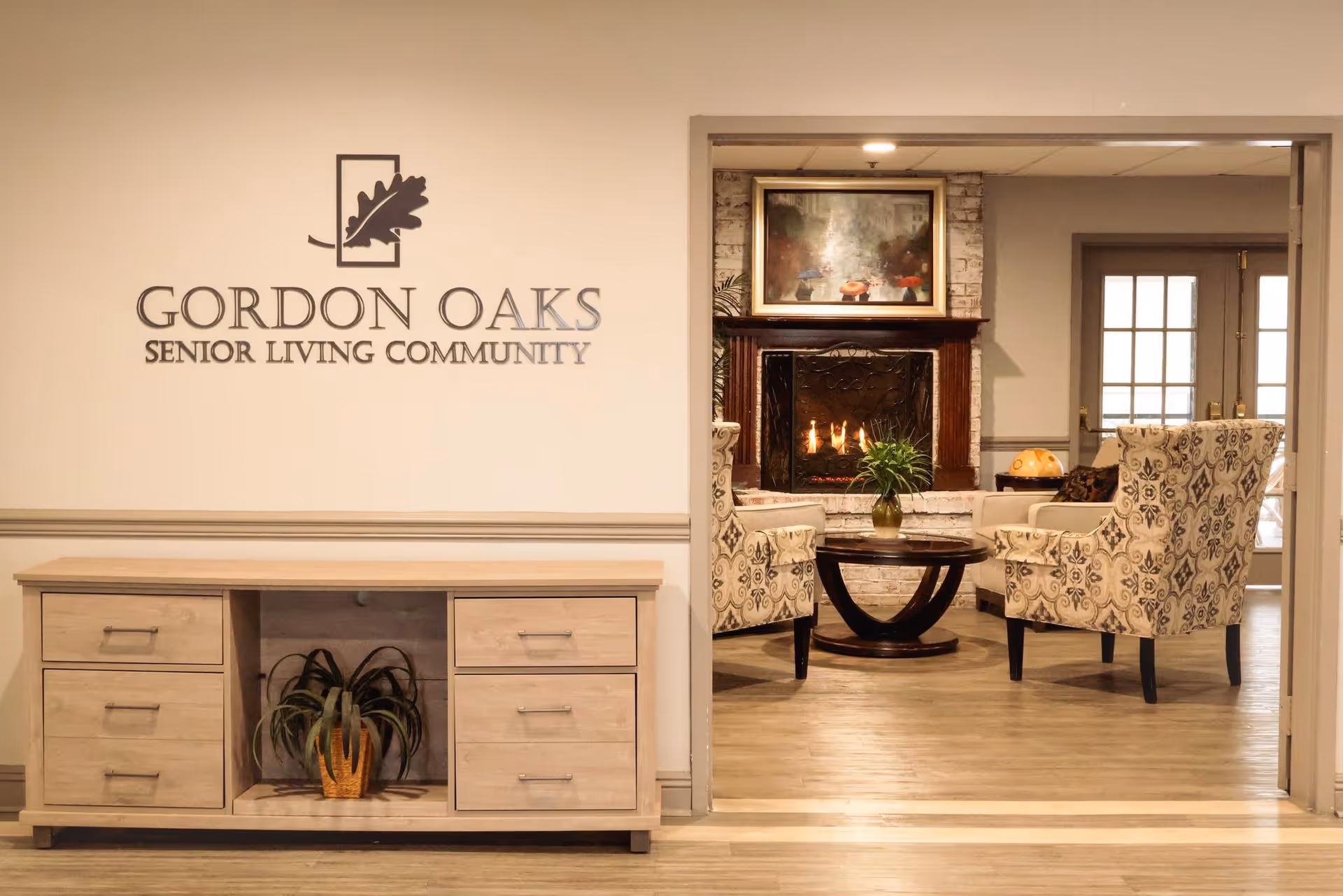 Interior view of Gordon Oaks Senior Living Community featuring a light wooden cabinet with a plant underneath and the community's logo on the wall above it. Through an open doorway, a cozy sitting area is visible with two patterned armchairs, a round wooden coffee table with a plant, and a lit fireplace with a painting above it.