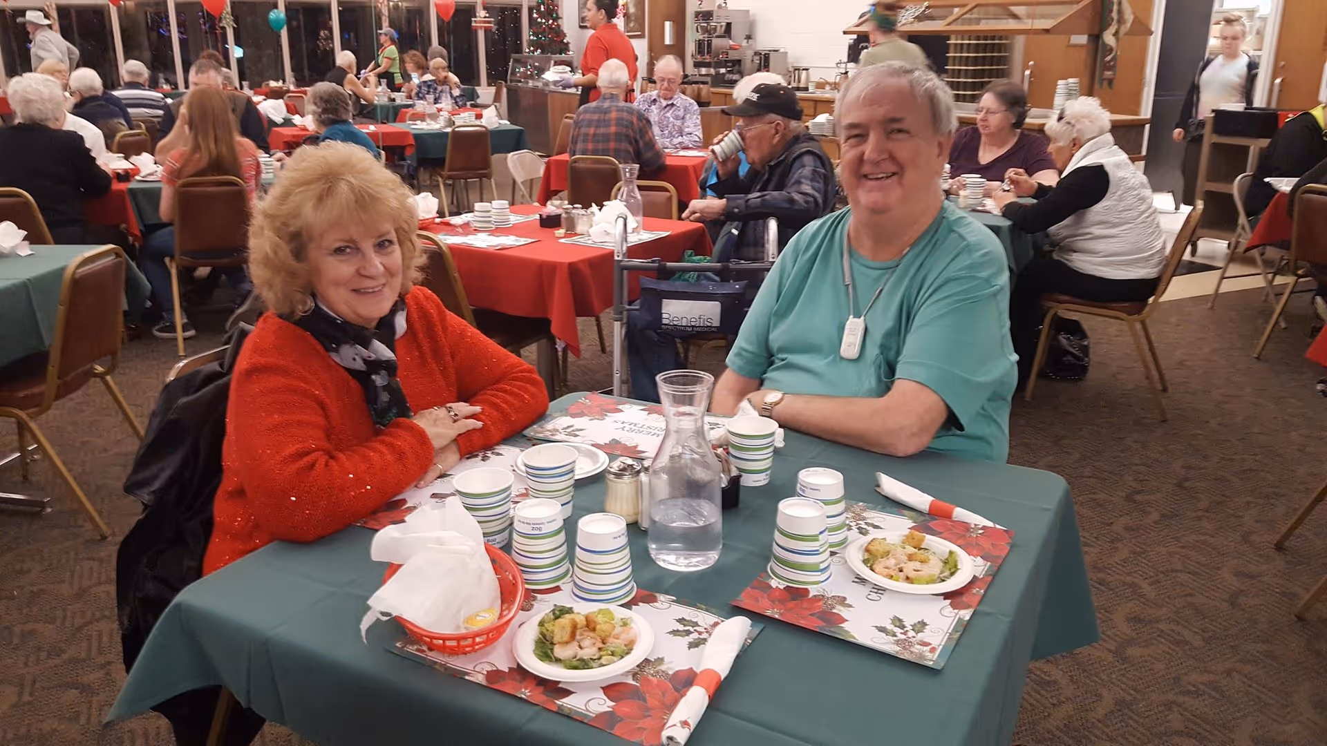 Two elderly people sitting at a table covered with a green tablecloth and holiday-themed placemats, smiling at the camera in a dining room filled with other seniors seated at tables with red and green tablecloths. Plates with food, cups, and a water pitcher are on the table. The room is decorated for the holidays with a Christmas tree visible in the background.