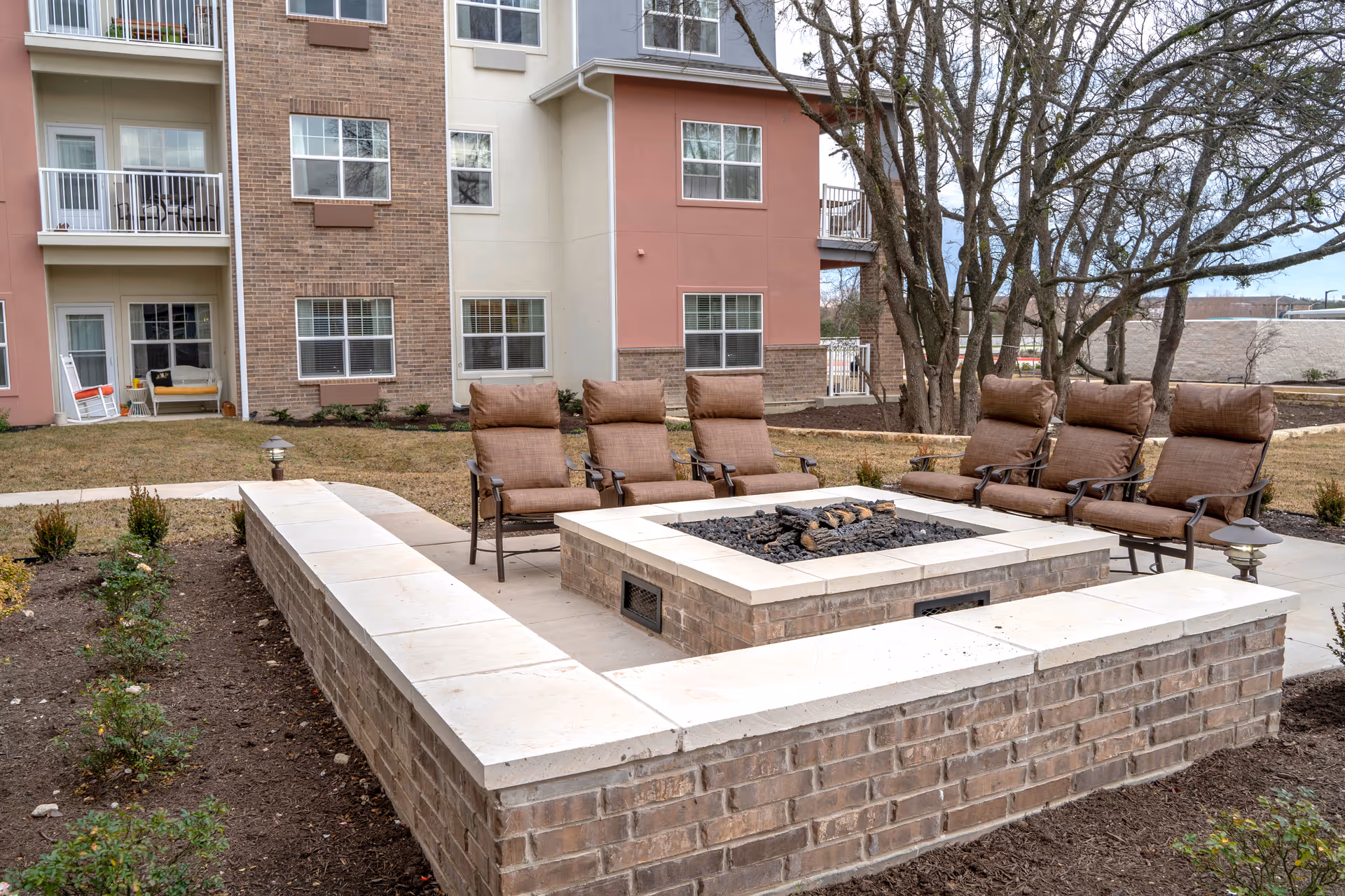 Outdoor seating area with six cushioned chairs arranged around a square brick fire pit. The area is part of a landscaped garden with small plants and trees, adjacent to a multi-story residential building with balconies and windows.